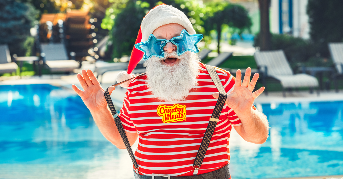 Santa Claus wearing sunglasses and a Country Meats shirt, standing by a pool with suspenders on